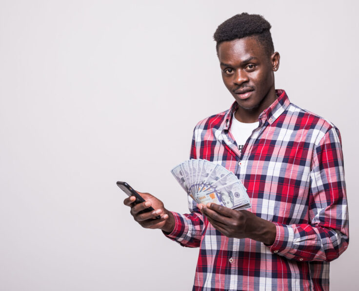 Portrait of satisfied young african man dressed in plaid shirt holding mobile phone and bunch of money banknotes isolated over white background