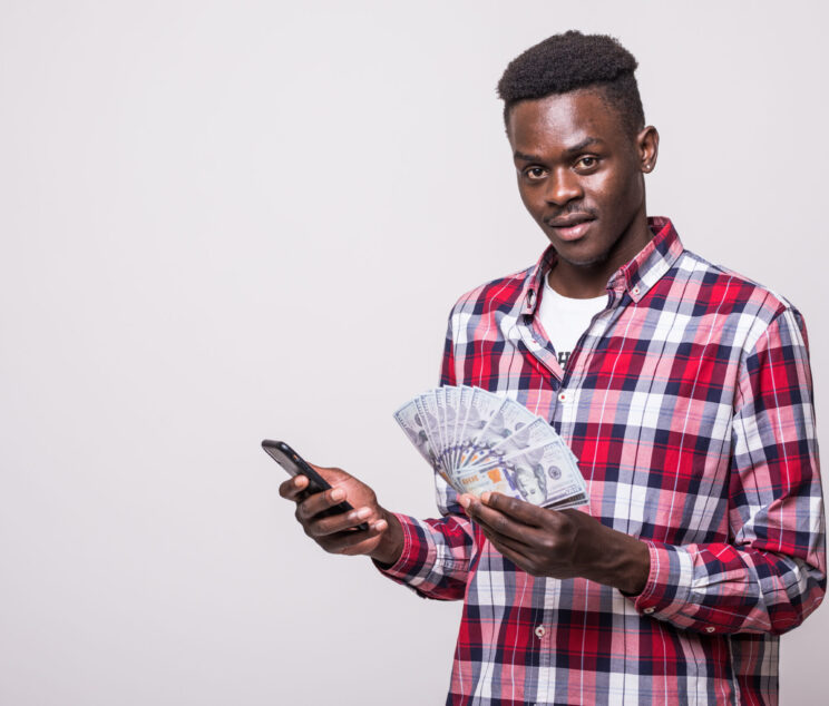 Portrait of satisfied young african man dressed in plaid shirt holding mobile phone and bunch of money banknotes isolated over white background